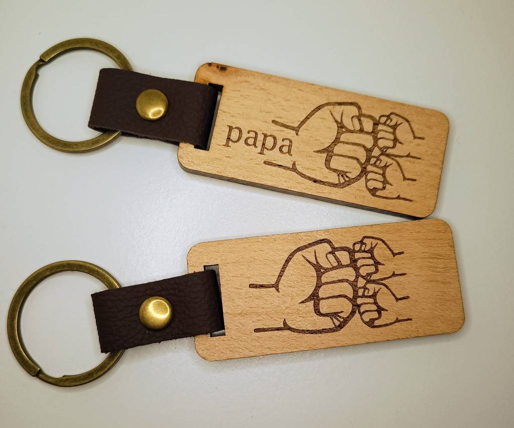 Deze foto toont een foto van twee houten sleutelhangers met een handgetekend beeld van een hand en de naam "papa".